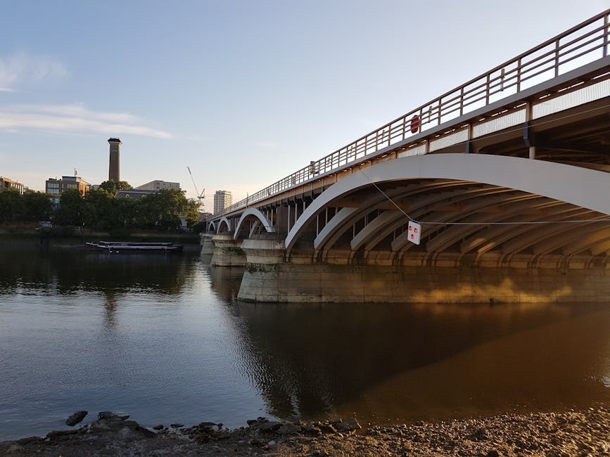 A large suspension bridge spanning a wide river under a partly cloudy sky, featuring tall white towers with red cables and railings. The bridge structure is supported by substantial concrete piers vis