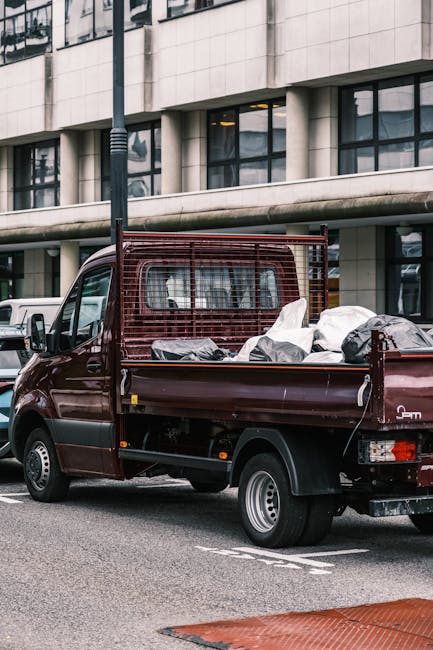 A burgundy flatbed van parked on a street outside a modern office building with large glass windows and concrete facade. The van's bed is loaded with various bulky waste items, including a white fabric-covered mattress, a wrapped sofa, and several large black and grey trash bags. A metal mesh safety barrier is installed at the head of the bed, and the vehicle's rear doors are open, indicating a loading or unloading process commonly associated with home relocation or furniture transport. Surrounding the van are other parked vehicles, and an overcast sky provides neutral lighting. The scene depicts a professional removals company's activity, likely involving the collection of large waste or furniture items during a house or office move, with the company's service focused on the safe and efficient removal of bulky waste, as referenced on the website manwithvanputney.co.uk.