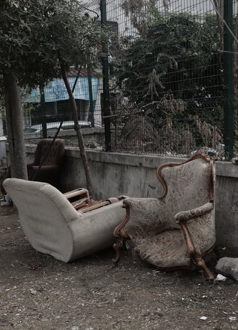 A photograph shows three old, upholstered armchairs and a sofa discarded outside on a dirt surface next to a concrete wall, with a metal fence and trees visible behind the fencing. The furniture appears worn, with fabric upholstery torn and faded, and wooden frame elements exposed or damaged. The chairs and sofa are positioned close together, some leaning against each other, indicating they are abandoned or awaiting disposal. The scene is outdoors with overcast lighting, and the ground has small rocks and debris scattered around. This image illustrates the disposal or removal of bulky household items, relevant to house removals or furniture transport services by companies such as Man with Van Putney, as seen on their website page about bulky waste removal in Putney.