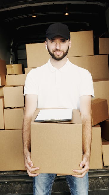 A man wearing a white polo shirt, black cap, and wireless earbuds stands inside the back of a van during a home relocation process, holding a single medium-sized cardboard box in front of him. The van's interior is filled with neatly stacked cardboard boxes of various sizes, some wrapped in plastic or packing paper, ready for transport. In the background, more boxes are visible toward the rear of the vehicle, suggesting an organized packing and loading process typical of moving services. The scene takes place outdoors, with the vehicle parked on a paved surface beneath a roof or overhang, with natural lighting illuminating the moving equipment and packed items. This image relates to professional removals and furniture transport, highlighting a typical step in packing and loading for house removals, as provided by Man with Van Putney.