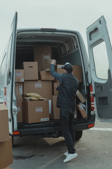 A man wearing a dark jacket, dark pants, white sneakers, and a cap is seen loading or unloading a variety of packed cardboard boxes into or from the back of a large grey van parked outside a property. The van's rear doors are wide open, revealing numerous boxes of different sizes, some with labels, stacked neatly inside. The boxes are wrapped in plastic or marked for easy identification, and some are placed on top of others to optimise space during the home relocation process. The scene appears to be set during daytime with natural light illuminating the loading area, which is adjacent to a paved driveway or street. Toward the front of the property, part of the entrance or doorway is visible, along with some outdoor ground surface. This image illustrates aspects of furniture transport, packing, and the loading process involved in professional removals services provided by Man with Van Putney, supporting last-minute or urgent move situations.