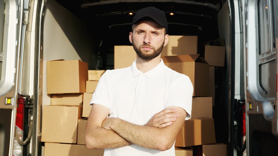 A man wearing a white polo shirt, black cap, and wireless earbuds stands inside the back of a van during a home relocation process, holding a single medium-sized cardboard box in front of him. The van's interior is filled with neatly stacked cardboard boxes of various sizes, some wrapped in plastic or packing paper, ready for transport. In the background, more boxes are visible toward the rear of the vehicle, suggesting an organized packing and loading process typical of moving services. The scene takes place outdoors, with the vehicle parked on a paved surface beneath a roof or overhang, with natural lighting illuminating the moving equipment and packed items. This image relates to professional removals and furniture transport, highlighting a typical step in packing and loading for house removals, as provided by Man with Van Putney.