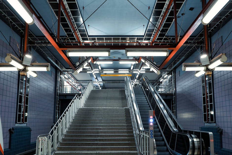 Interior view of an urban underground station stairwell with a wide concrete staircase and an adjoining escalator. The stairs are equipped with metal handrails on both sides, and the escalator has a glass and metal railing. Surrounding walls are tiled in blue, and the ceiling features a symmetrical grid of exposed structural beams painted in orange and black. Overhead, lighting fixtures provide bright illumination, with additional wall-mounted lights on either side of the staircase. Visible equipment includes a small sensor or information panel at the base of the escalator. This environment is a typical setting for home relocation logistics, where furniture and packed boxes may be transported via stairs or escalator, supported by removal specialists such as Man with Van Putney to facilitate efficient moving services within urban transit spaces.