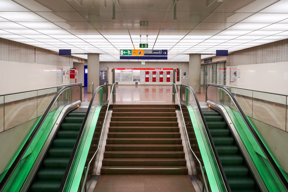 Interior view of an urban underground station stairwell with a wide concrete staircase and an adjoining escalator. The stairs are equipped with metal handrails on both sides, and the escalator has a glass and metal railing. Surrounding walls are tiled in blue, and the ceiling features a symmetrical grid of exposed structural beams painted in orange and black. Overhead, lighting fixtures provide bright illumination, with additional wall-mounted lights on either side of the staircase. Visible equipment includes a small sensor or information panel at the base of the escalator. This environment is a typical setting for home relocation logistics, where furniture and packed boxes may be transported via stairs or escalator, supported by removal specialists such as Man with Van Putney to facilitate efficient moving services within urban transit spaces.