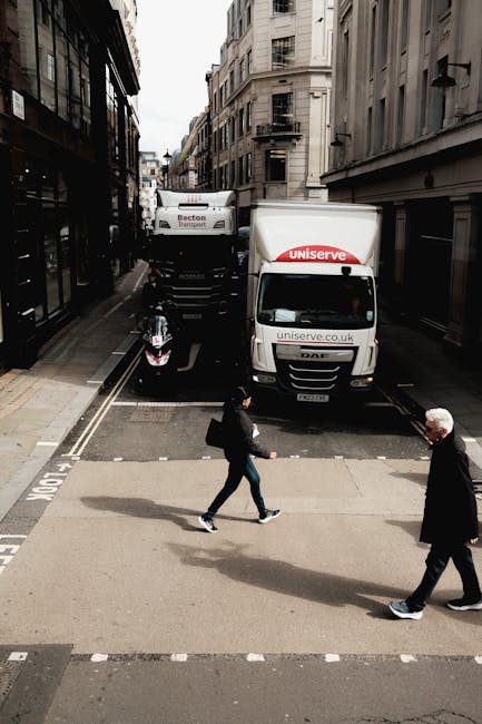 A street scene in Putney featuring a small residential road with tall, multi-storey buildings on both sides. In the foreground, two pedestrians are crossing the road—one person dressed in dark clothing carrying a backpack and the other with white hair wearing a dark coat. Behind them, a parked motorcycle is visible alongside a white box truck with the label 'Uniserve' on the front and side panels, positioned partially on the pedestrian crossing area. Further down the street, a larger white van with the branding 'Becton Transport' is parked behind the truck, with narrow alleyways and additional building facades extending into the background. The scene is captured during daylight with overcast weather, and the street appears to be quiet, with no other moving vehicles or pedestrians visible. This image relates to home relocation and furniture transport services, showcasing the typical urban environment in which [COMPANY_NAME] might operate for house removals or moving logistics.