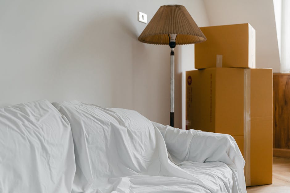 Interior scene of a living room with a white fabric-covered sofa positioned against a light-colored wall. To the right of the sofa, there are three stacked cardboard moving boxes, two large and one smaller, all sealed with packing tape, indicating preparations for a home relocation. A floor-standing lamp with a brown pleated lampshade is situated behind the boxes, providing ambient lighting. The room appears well-lit with natural light coming from a window on the right side, partially visible with a sheer curtain. The environment suggests a space in the process of being packed or prepared for搬家, with the visible items and setup reflecting common elements involved in furniture transport and packing during house removals, as provided by [COMPANY_NAME].