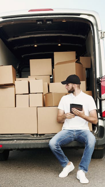 A man wearing a white t-shirt, blue jeans, white sneakers, and a black cap is sitting on the ground beside an open moving van, which is filled with neatly stacked cardboard boxes of various sizes. The boxes are secured with plastic wrap and are placed on the vehicle's floor and platform, indicating a home relocation process. The man is holding a tablet device and appears to be reviewing or managing the loading and packing process. The inside of the van shows a dark ceiling and is positioned on a paved surface, with part of a residential or commercial setting visible in the background. This image reflects professional packing, furniture transport, and loading activities associated with house removals, relevant to services offered by Man with Van Putney as part of their removal solutions.