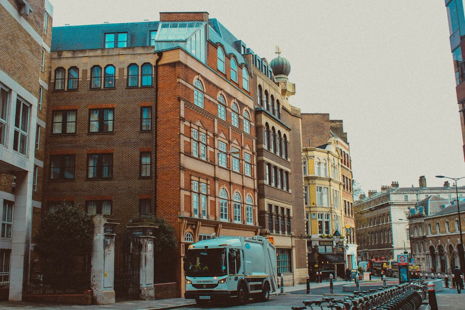 The image shows a city street scene with a white van, likely used for house removals or moving services, parked in front of a row of historic and modern buildings with varied architectural styles and brick facades. The van is positioned on the road, close to the pavement, with its front facing towards the right side of the frame. The setting appears to be daytime under natural light, with overcast sky overhead. In the background, additional vehicles and urban infrastructure are visible, indicating an active relocation or furniture transport environment. Nearby, a small section of bicycles or racks for bicycle parking can be seen on the right side, along with street furniture and bollards. [COMPANY_NAME] provides professional removals in this area, supporting home relocation and furniture transport by facilitating the loading and unloading process against a backdrop of city buildings, emphasizing the logistics involved in urban house removals and packing and moving procedures.