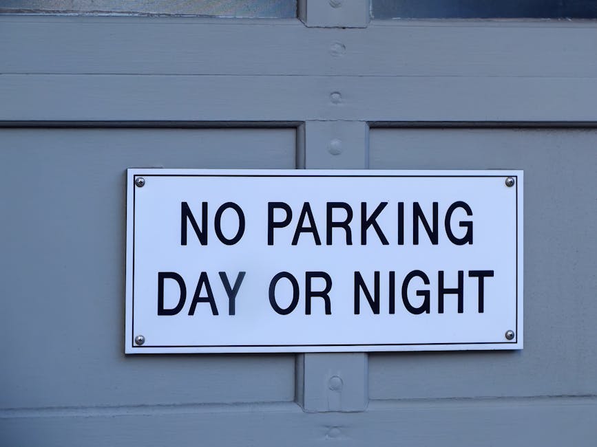 A rectangular white sign with bold black lettering that reads 'NO PARKING DAY OR NIGHT' affixed to a light grey wooden door or wall. The sign is attached with four visible screws, one in each corner. The surface behind the sign is made of horizontal wooden panels with a slight texture, and the area appears to be outdoors or at the exterior of a building. This signage relates to parking regulations, which are relevant to loading bay rules during home relocation or furniture transport activities. The setting is well-lit, and the focus is on the sign, emphasizing its importance for vehicle restrictions in the vicinity. Man with Van Putney, a professional removals service provider, would be aware of such signage when planning residential or commercial moves to ensure compliance with local loading and parking regulations.