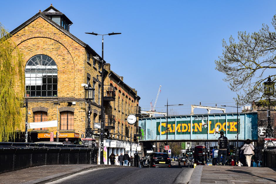 The image depicts a busy street scene in Putney with a large, historic brick building on the left featuring arched windows and a clock mounted on a pole. Several pedestrians are walking along the pavement, some near parked cars and others near storefronts, including one with an orange sign for orange juice. In the background, there is a large railway bridge with a blue container marked 'CADDIN LOCK' loaded on a train passing overhead, and visible construction cranes further in the distance. The scene is well-lit with clear, blue sky overhead and a few trees with budding leaves on the right. The street appears to be the route for home relocation activities, with the presence of vehicles and street-level activity indicating the movement of furniture and packing materials, which aligns with house removals and furniture transport services provided by Man with Van Putney. This setting reflects a typical urban environment where careful loading, unloading, and logistics are integral to effective moving and relocation services.