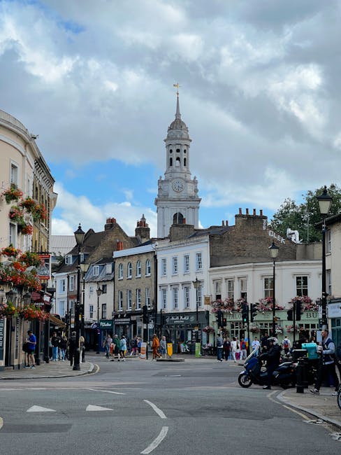 A busy street scene in Putney featuring a mix of historic and modern buildings, with shopfronts and pedestrians on the pavement. In the background, a tall white clock tower with a golden weather vane at the top rises above the surrounding structures. Several shops display signage, and hanging flower baskets decorate the streetscape. Vehicles, including motorcycles and cars, are visible on the road, while people walk along the sidewalks. Overhead, a partly cloudy sky provides natural lighting. This setting illustrates an urban environment where house removals or furniture transport could be taking place, with visible activity relevant to home relocation and professional moving services, supported by the presence of the environment typical for loading and unloading processes in a central Putney location, as seen in the context of [PAGE_TITLE].