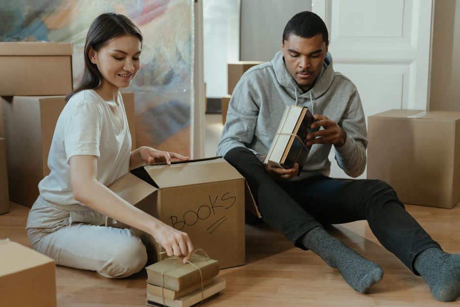 A person with light skin is seen loading a small stack of books into an open cardboard moving box, which is positioned on a plain white surface. The books have hard covers in neutral tones, some with 