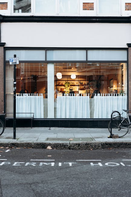 A clear, large window of a ground-floor commercial property reveals the interior of a well-lit cafe or restaurant. Inside, wooden shelves stocked with various items are visible against a wall, with spherical pendant lights hanging from the ceiling. The front of the establishment is separated from the sidewalk by light blue sheer curtains, partially drawn back. Outside, the pavement includes a bicycle rack with one black bicycle secured and a second bicycle leaning nearby, and a designated parking space marked 'PERMIT' on the road. The building’s facade features a white and black exterior, with a small blue parking sign attached near the entrance. The overall scene suggests an urban setting with a focus on the storefront, with no visible furniture or moving activity, and the environment appears quiet and well-maintained, consistent with a typical interior of a café or restaurant in an area that may require parking permits for vehicle access. Man with Van Putney may assist in local home or business relocations involving such premises.