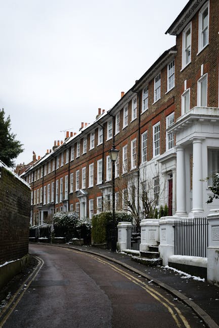A row of traditional red brick terraced houses with white window frames and decorative white columns at the entrances lines a narrow, curving residential street in Putney during winter. The street is paved with asphalt and features a double yellow line along the edge, indicating parking restrictions. There are black wrought iron lampposts and small gardens with shrubbery and trees, some of which are leafless, suggesting the colder season. In the foreground, a loading area or driveway adjacent to one of the properties is visible, with a few cardboard boxes, wrapped furniture, and packing materials placed near the doorway, indicating home relocation preparations. A Man with Van Putney vehicle is partially visible, parked on the street, with a person lifting a large, wrapped piece of furniture onto a trolley nearby, assisting with furniture transport as part of the packing and moving process. The scene captures a typical residential move involving careful handling of household items in a quiet London neighbourhood, aligned with professional removals services and parking permit considerations for house removals in this area.
